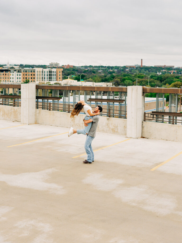 A man and a woman in a rooftop parking garage in San Antonio. The man is wearing a grey t-shirt and jeans, and the woman is wearing a white shirt and ripped jeans. The photo showcases the scenery of San Antonio. The man is holding the woman around her butt, and is spinning her in a circle. The woman is leaning back slightly.