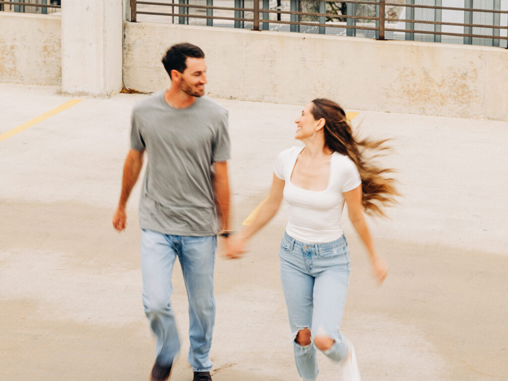 A man and a woman in a rooftop parking garage in San Antonio. The man is wearing a grey t-shirt and jeans, and the woman is wearing a white shirt and ripped jeans. The man and woman are holding hands and looking at each other as they run through the garage.