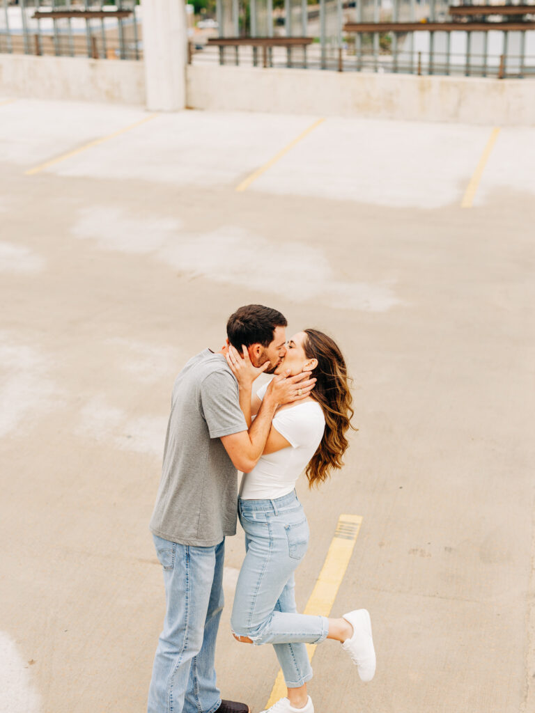A man and a woman in a rooftop parking garage in San Antonio. The man is wearing a grey t-shirt and jeans, and the woman is wearing a white shirt and ripped jeans. The couple is engaged in a passionate kiss, and have their hands on each other's faces.