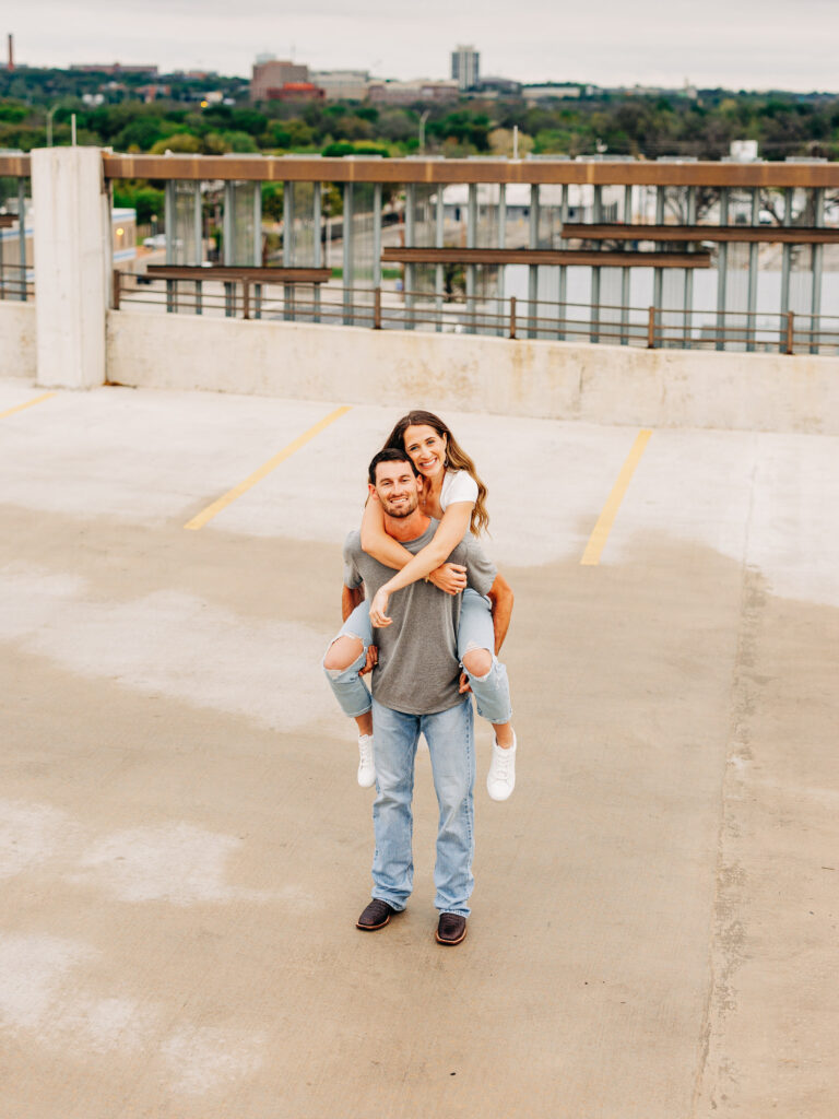 A man and a woman in a rooftop parking garage in San Antonio. The man is wearing a grey t-shirt and jeans, and the woman is wearing a white shirt and ripped jeans. The man is holding the woman on his back, and is supporting her by grasping under her knees. The woman has her arms wrapped loosely around his neck.
