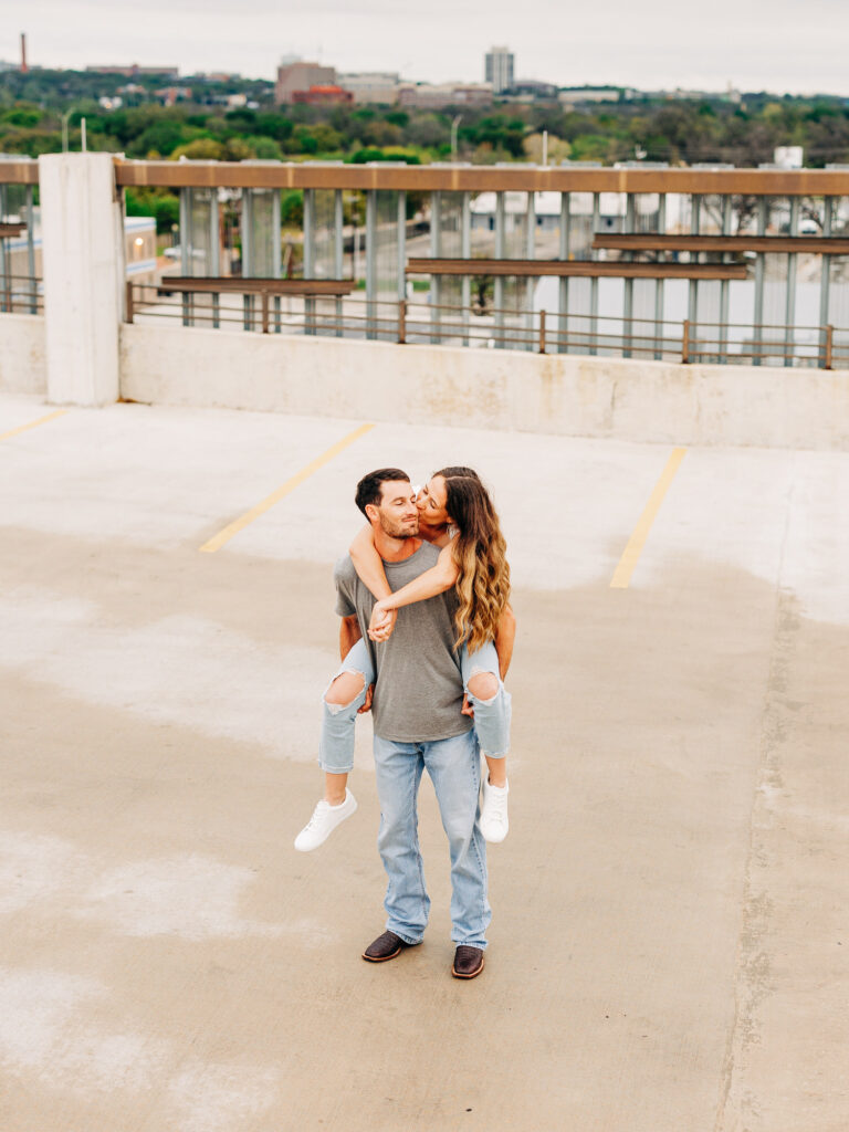A man and a woman in a rooftop parking garage in San Antonio. The man is wearing a grey t-shirt and jeans, and the woman is wearing a white shirt and ripped jeans. The man is holding the woman on his back, and is supporting her by grasping under her knees. The woman has her arms wrapped loosely around his neck. The woman is kissing the man's cheek.