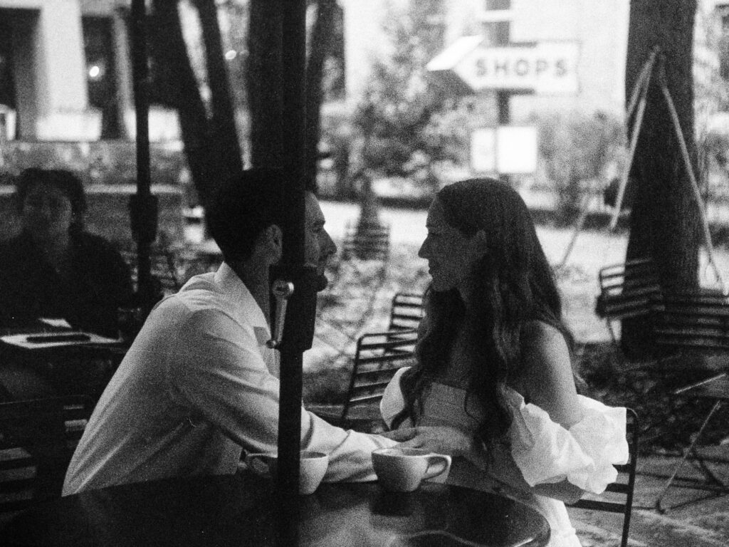 A monochrome film photograph of an engaged couple sitting at a patio table. There are two cups of coffee sitting on the table. The man is leaning into the woman and holding her hand.