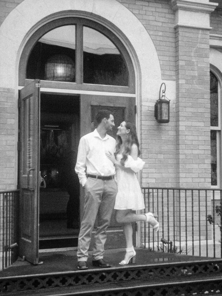 A monochrome film photograph of a man and a woman standing in front of a brick building. The man is looking down at the woman as she lovingly looks up at him while placing a hand on his chest.