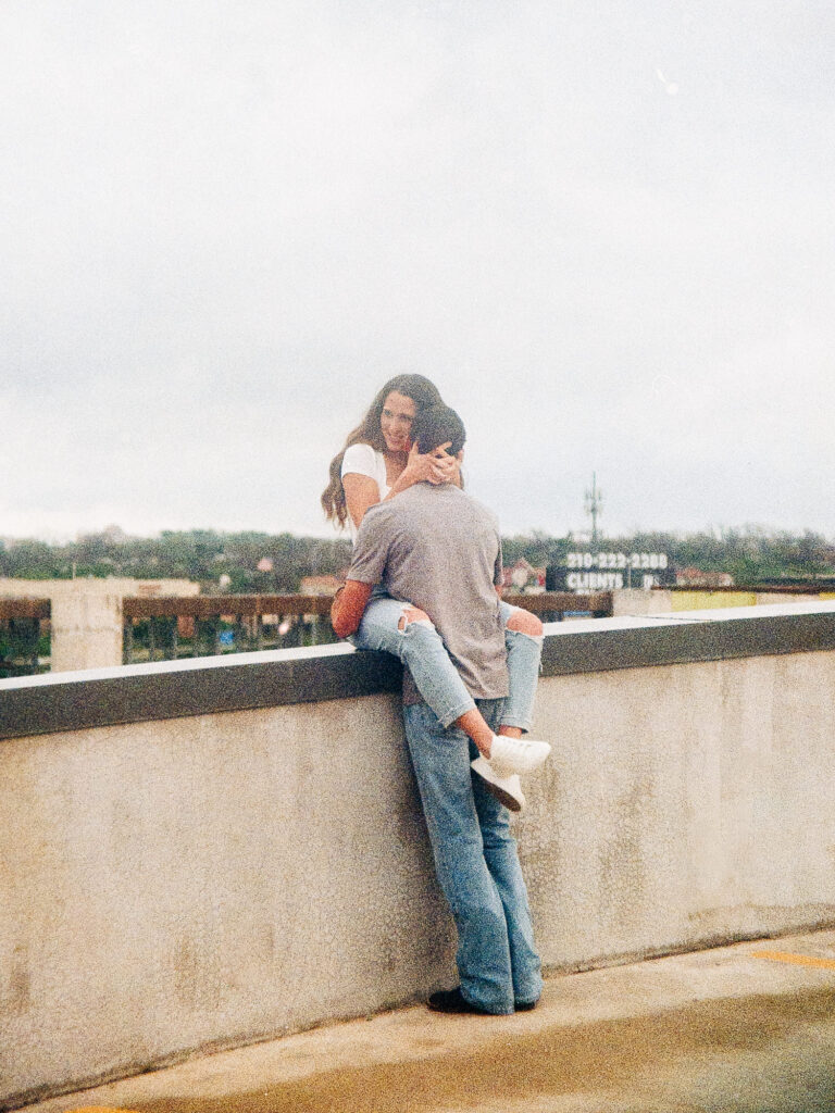 A film photo of an engaged couple in a rooftop parking garage. The background showcases downtown San Antonio. The woman is sitting on a railing, wrapping her legs around the man and her arms around his neck. The man is holding onto her.