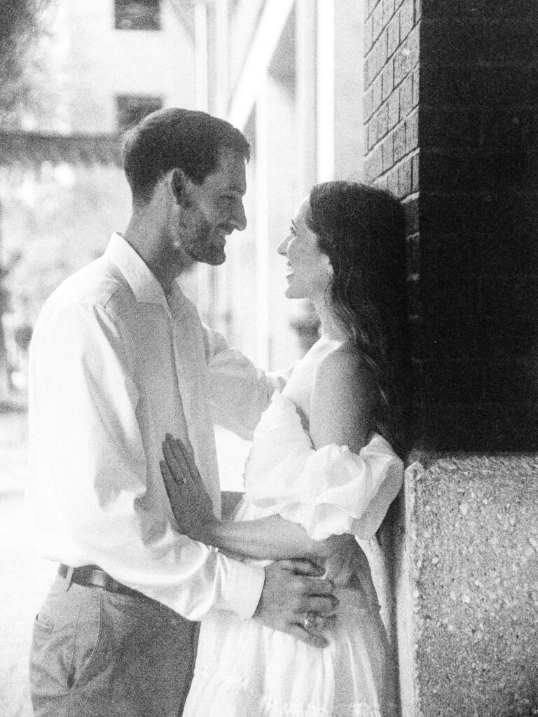 A monochrome photograph taken on film of an engaged couple. The man is leaning the woman against a brick wall and holding her waist.