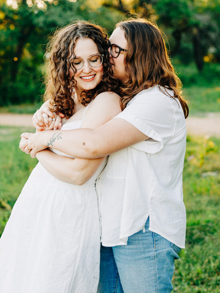 A photo of two engaged women holding each other in a park in Austin, TX. One woman is wearing a white spaghetti strap dress and the other is wearing a white button up top and blue jeans. There are trees with green leaves in the background. One woman is kissing the other's head