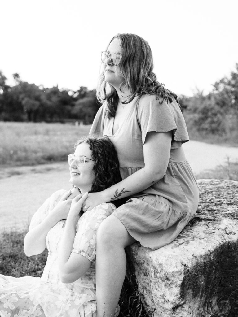 A monochrome photo of an LGBTQ+ engaged couple. One of them is sitting on a rock and the other is sitting on the ground in between the other's legs. They are holding hands and looking into the distance.