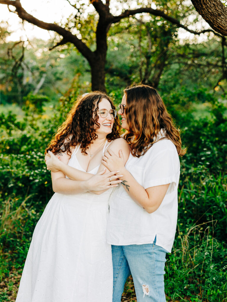 A photo of two engaged women holding each other in a park in Austin, TX. One woman is wearing a white spaghetti strap dress and the other is wearing a white button up top and blue jeans. There are trees with green leaves in the background.