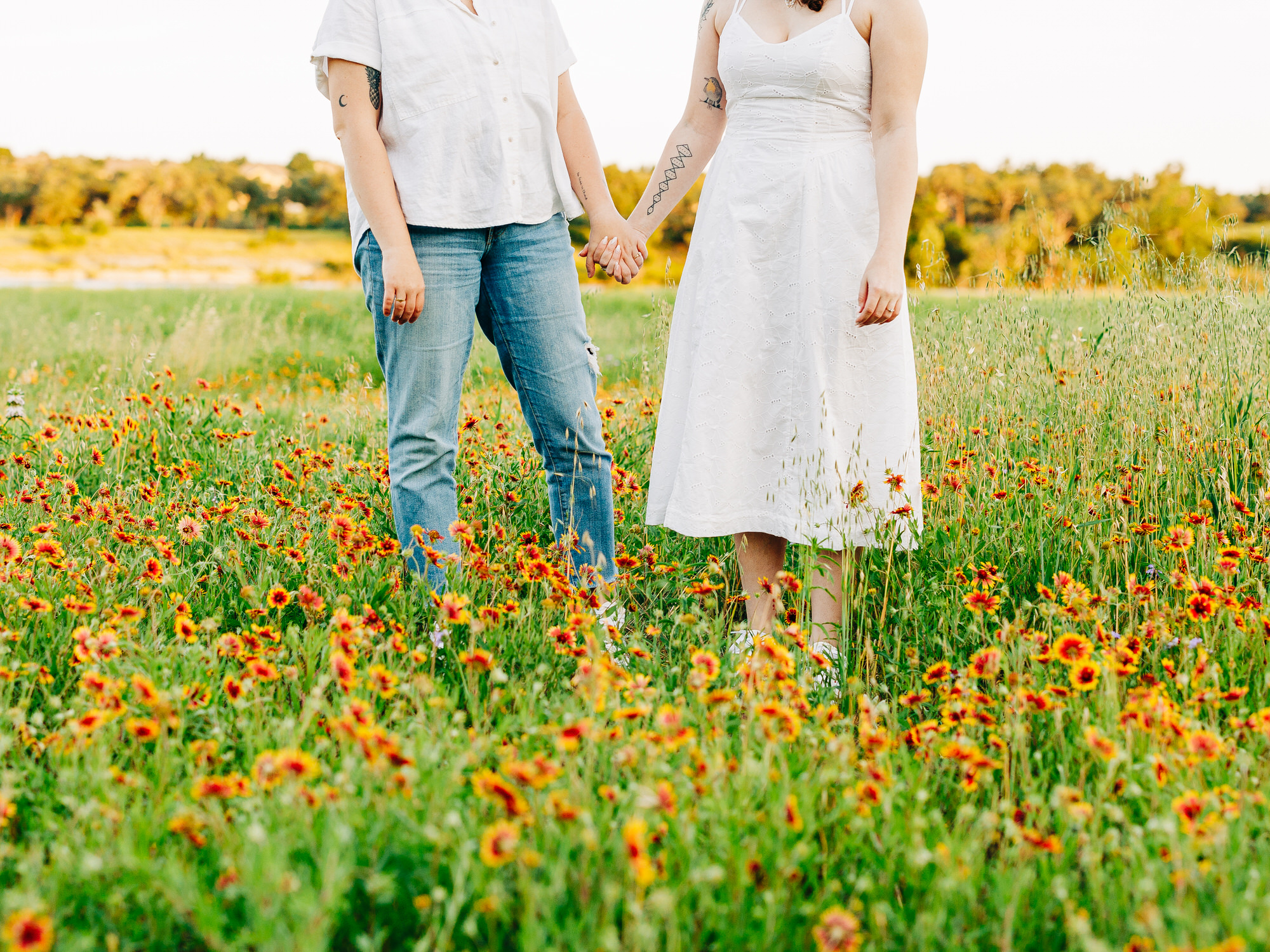 A photo of two engaged women standing in a field of wildflowers in Austin, TX. The flowers are yellow with a red center. One of the women is wearing a white spaghetti strap dress and the other is wearing a white button-up shirt and ripped blue jeans. The women are holding hands.