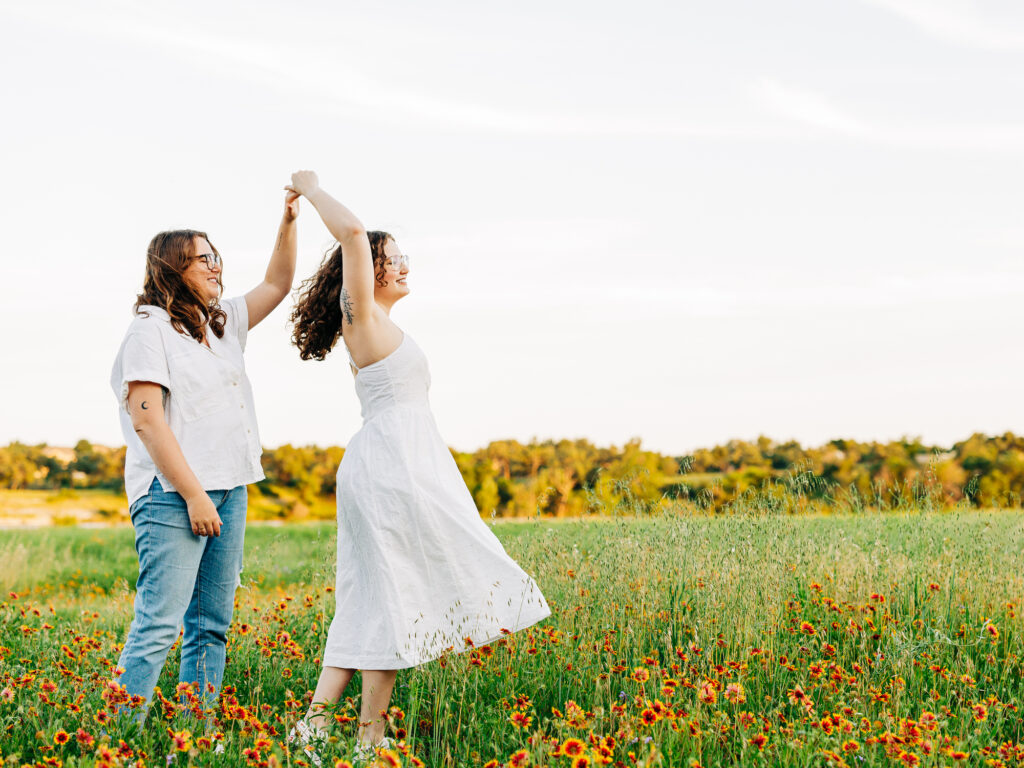 A photo of two engaged women standing in a field of wildflowers in Austin, TX. The flowers are yellow with a red center. One of the women is wearing a white spaghetti strap dress and the other is wearing a white button-up shirt and ripped blue jeans. The women are dancing and smiling.