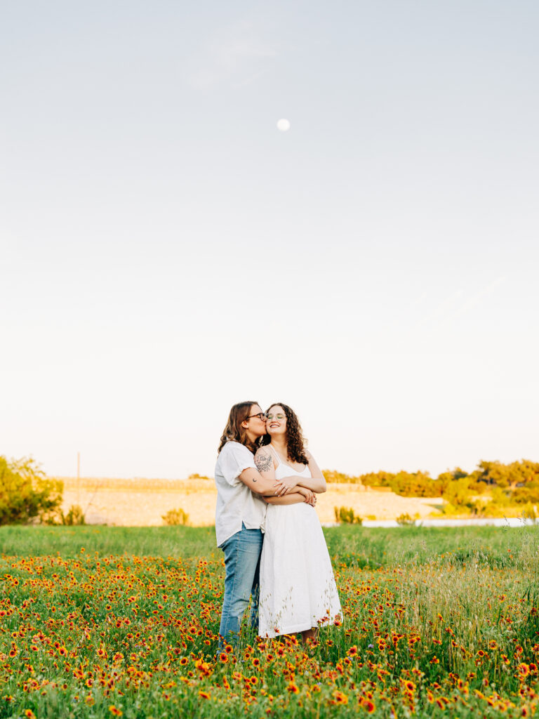 A photo of two engaged women standing in a field of wildflowers in Austin, TX. The flowers are yellow with a red center. One of the women is wearing a white spaghetti strap dress and the other is wearing a white button-up shirt and ripped blue jeans. The women are holding each other and looking at each other. The moon is a full moon and is visible in the sky.