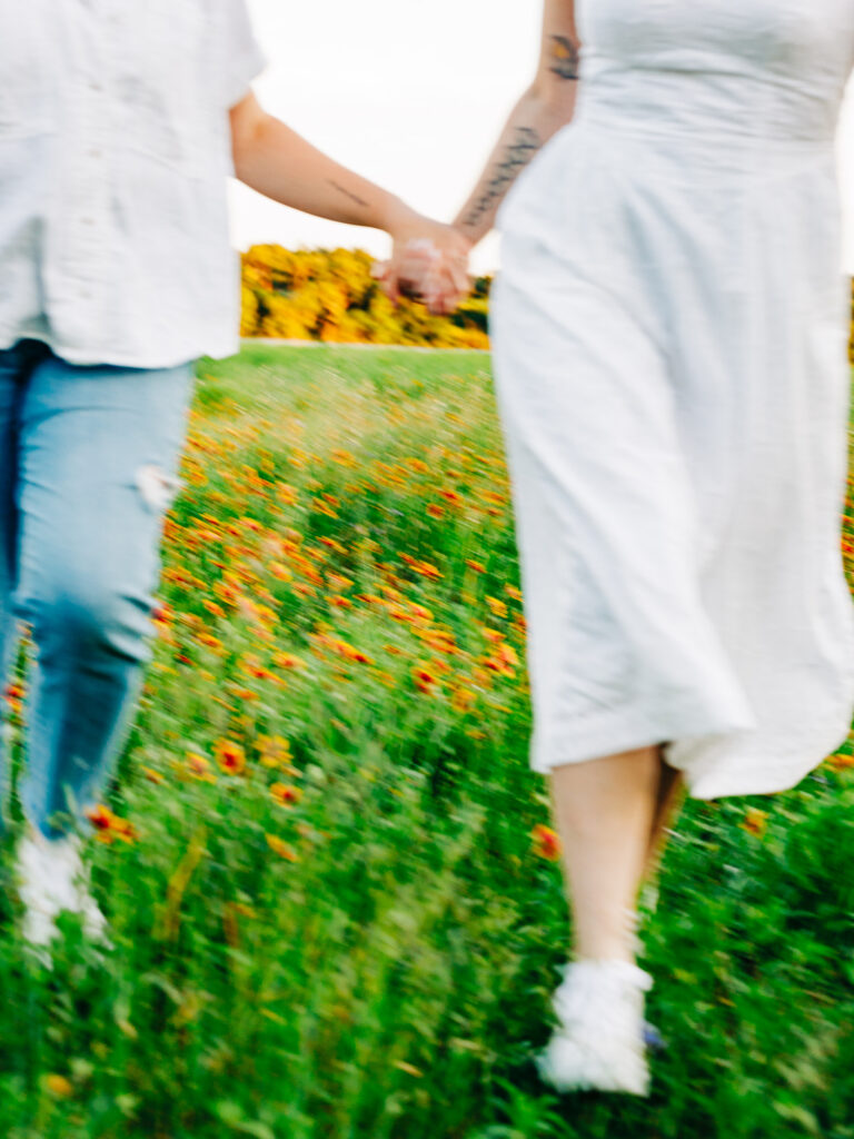 A photo of two engaged women in a field of wildflowers in Austin, TX. The flowers are yellow with a red center. One of the women is wearing a white spaghetti strap dress and the other is wearing a white button-up shirt and ripped blue jeans. The women are running towards the camera and the image is blurry.