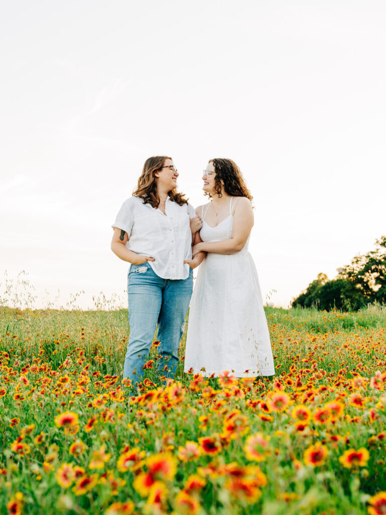 A photo of two engaged women standing in a field of wildflowers in Austin, TX. The flowers are yellow with a red center. One of the women is wearing a white spaghetti strap dress and the other is wearing a white button-up shirt and ripped blue jeans. The women are holding each other and looking at each other.