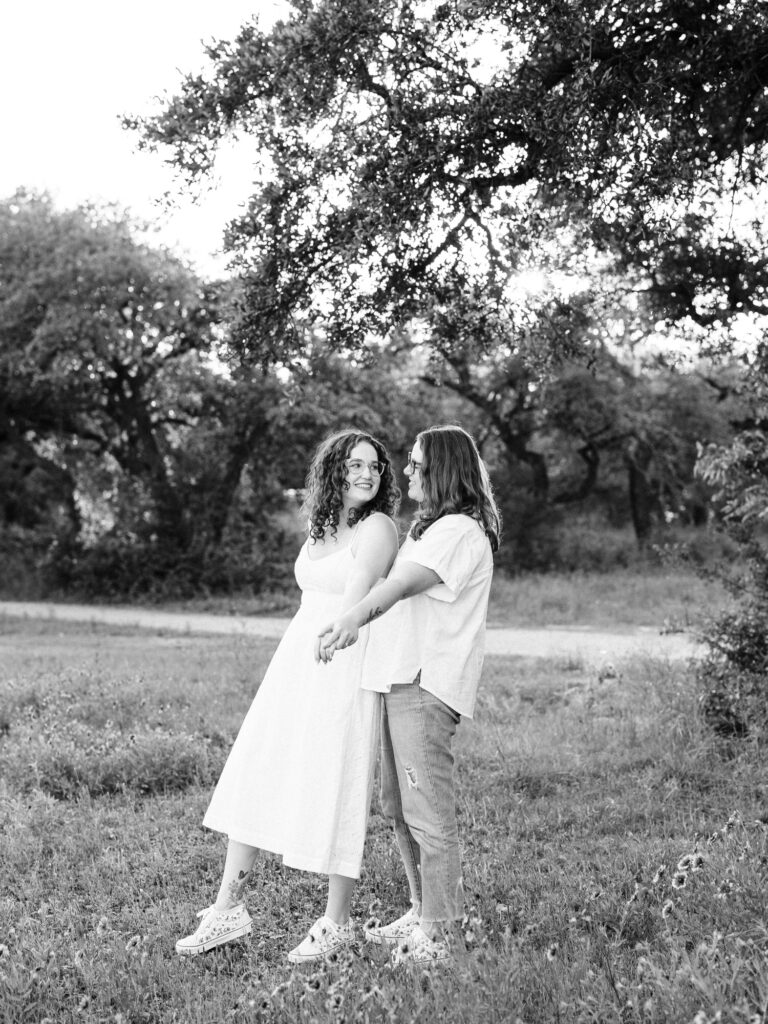 A monochrome image of two engaged women standing in a park in Austin, TX. One woman is leaning into the other as they hold hands, both facing the same direction. They are looking at eachother. One woman is wearing a white spaghetti strap dress and the other is wearing a white button up and blue jeans.
