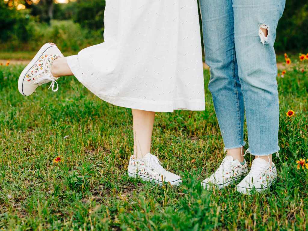 Two engaged women standing in the grass wearing matching Vans shoes. The shoes are beige and contain multi-colored florals on the sides.