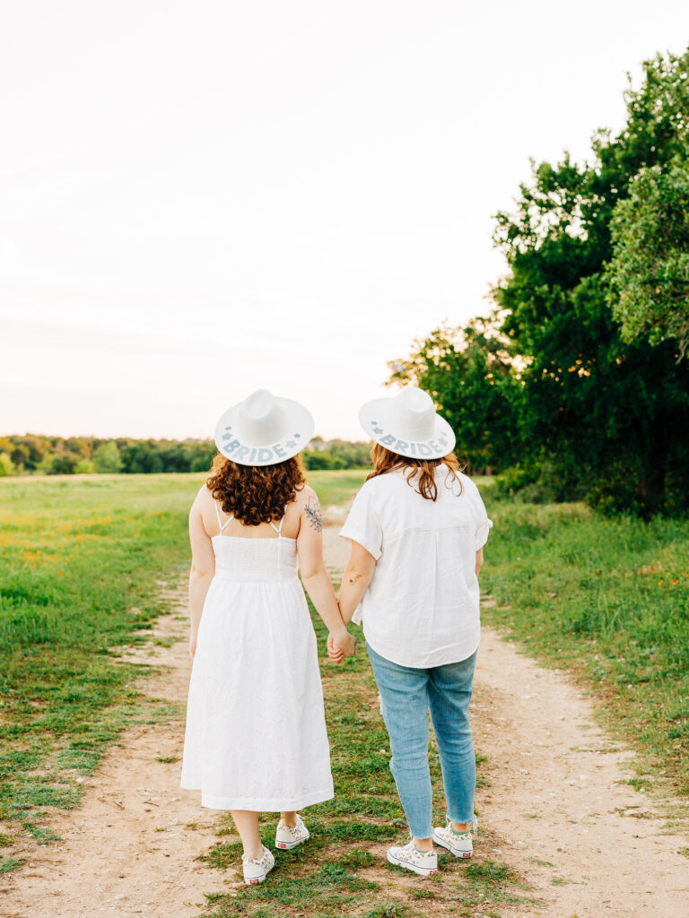 A photo of an engaged couple standing on a path at a park in Austin, Texas. Both women are wearing hats that say "Bride". They are holding hands and walking away from the camera. One is wearing a white spaghetti strap dress and the other is wearing a white button up shirt and ripped jeans.