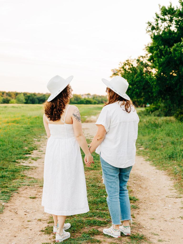 A photo of an engaged couple standing on a path at a park in Austin, Texas. Both women are wearing hats that say "Bride". They are holding hands and walking away from the camera. One is wearing a white spaghetti strap dress and the other is wearing a white button up shirt and ripped jeans.