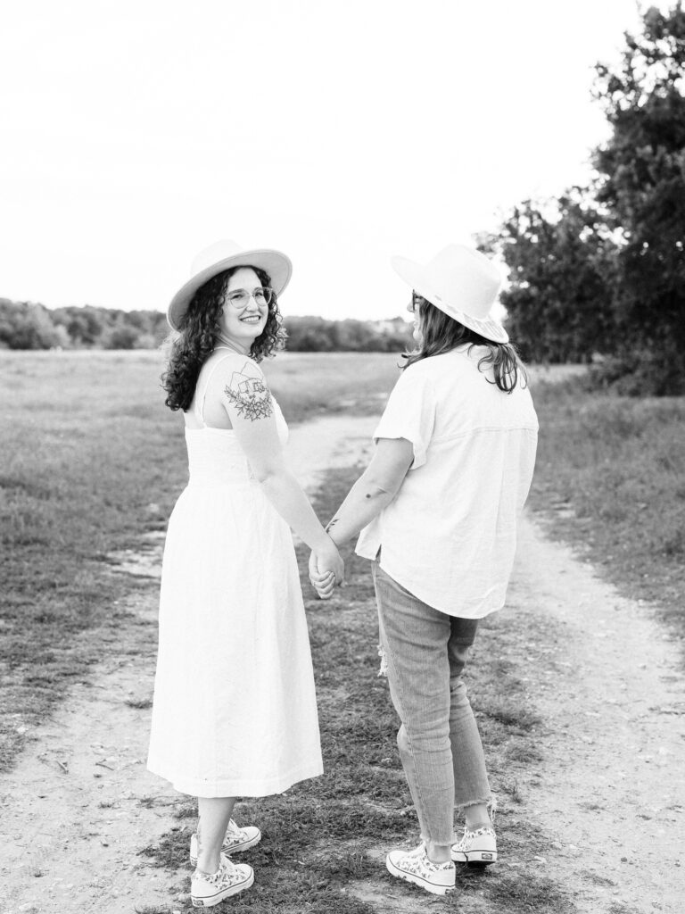 A monochrome photo of an engaged couple standing on a path at a park in Austin, Texas. Both women are wearing hats that say "Bride". They are holding hands and walking away from the camera as one of them looks back. One is wearing a white spaghetti strap dress and the other is wearing a white button up shirt and ripped jeans.