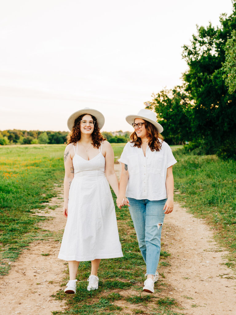 A photo of an engaged couple standing on a path at a park in Austin, Texas. Both women are wearing hats that say "Bride". They are holding hands and walking towards the camera as one of them looks at the camera. One is wearing a white spaghetti strap dress and the other is wearing a white button up shirt and ripped jeans.