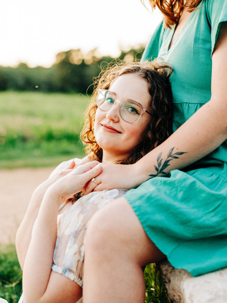 A photo of an LGBTQ+ engaged couple. One of them is sitting on a rock and the other is sitting on the ground in between the other's legs. They are holding hands. The woman sitting on the ground is looking towards the camera.
