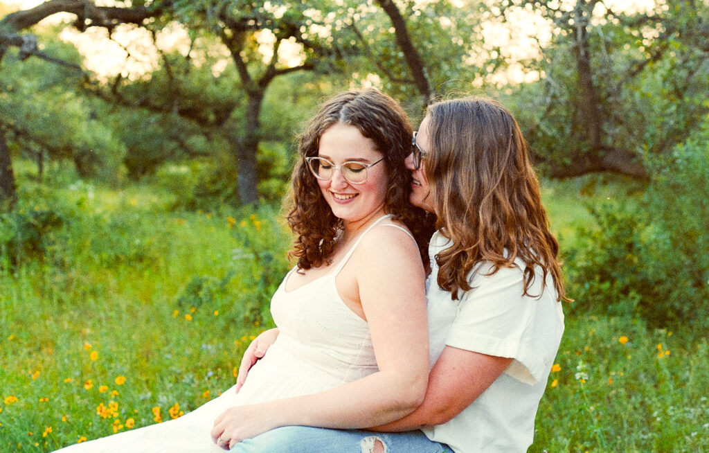 Two engaged women sitting in a grassy field in Austin, Texas, while one of them whispers in the other's ear. One is wearing a white dress and the other is wearing a white button up top and ripped jeans