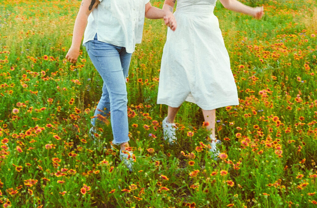 two soon to be brides running through a field of flowers in Austin, Texas during their engagement photography session. taken on film