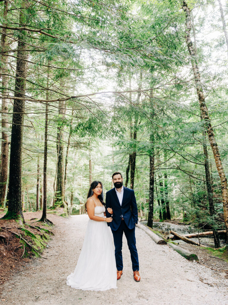 A couple standing on a forest path, surrounded by tall trees. The woman is in a strapless white dress, and the man wears a dark suit with brown shoes. Both look serene, holding hands, with soft sunlight filtering through the trees.