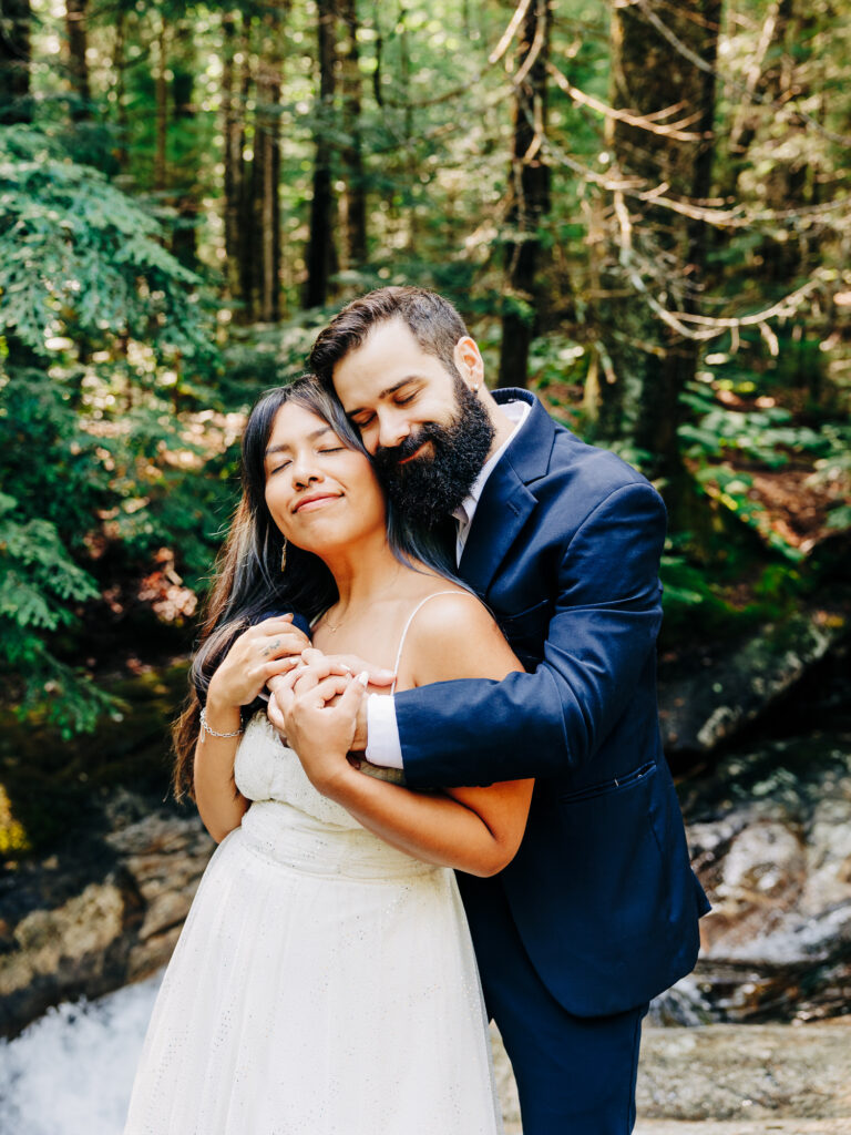 The man tenderly wraps his arms around the woman, standing close to a rushing forest stream. Both are relaxed, the man resting his head gently on hers, while she smiles with her eyes closed, enjoying the embrace.