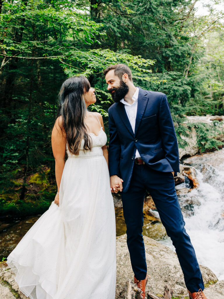 The couple standing hand in hand on a rock near a waterfall, surrounded by lush greenery. The man gazes lovingly at the woman, who looks back at him with a smile. The woman's white dress contrasts beautifully with the natural forest setting.