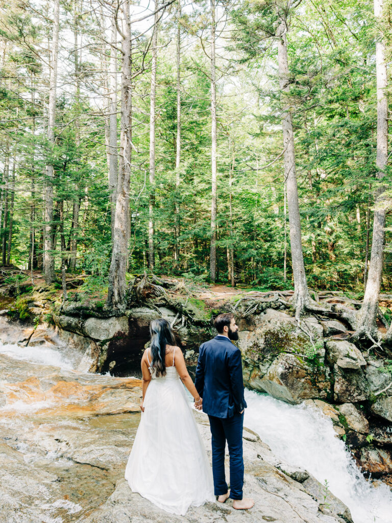 The couple stands on a rocky ledge, overlooking a cascading waterfall surrounded by towering trees. The woman’s white dress flows elegantly, and the man, barefoot in his suit, stands beside her as they take in the peaceful, natural beauty of Franconia Notch.