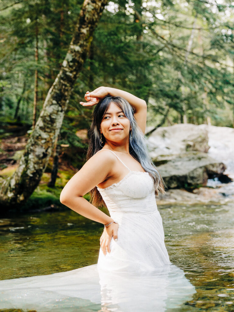 The woman, in a white dress, poses confidently in a shallow creek, surrounded by trees. She has one hand on her hip and the other raised above her head, with a soft smile as she stands in the clear water.