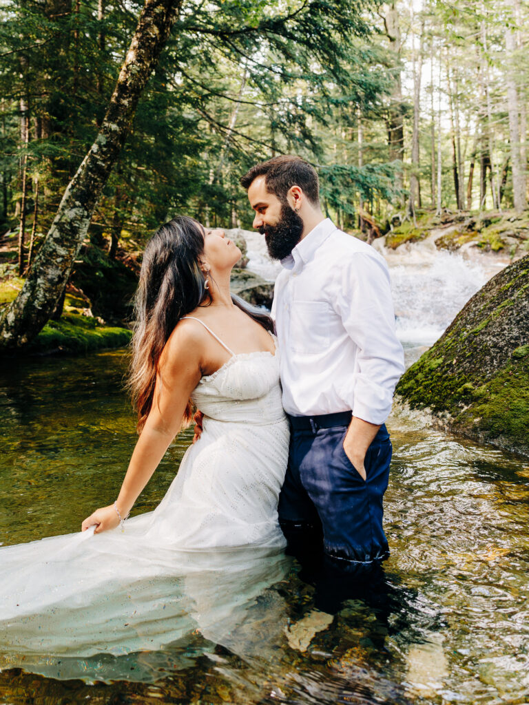 The couple stands in a shallow creek with their lower halves submerged in water. The woman in a white dress leans back, while the man in rolled-up pants gazes down at her lovingly. A waterfall is visible in the background, surrounded by moss-covered rocks.