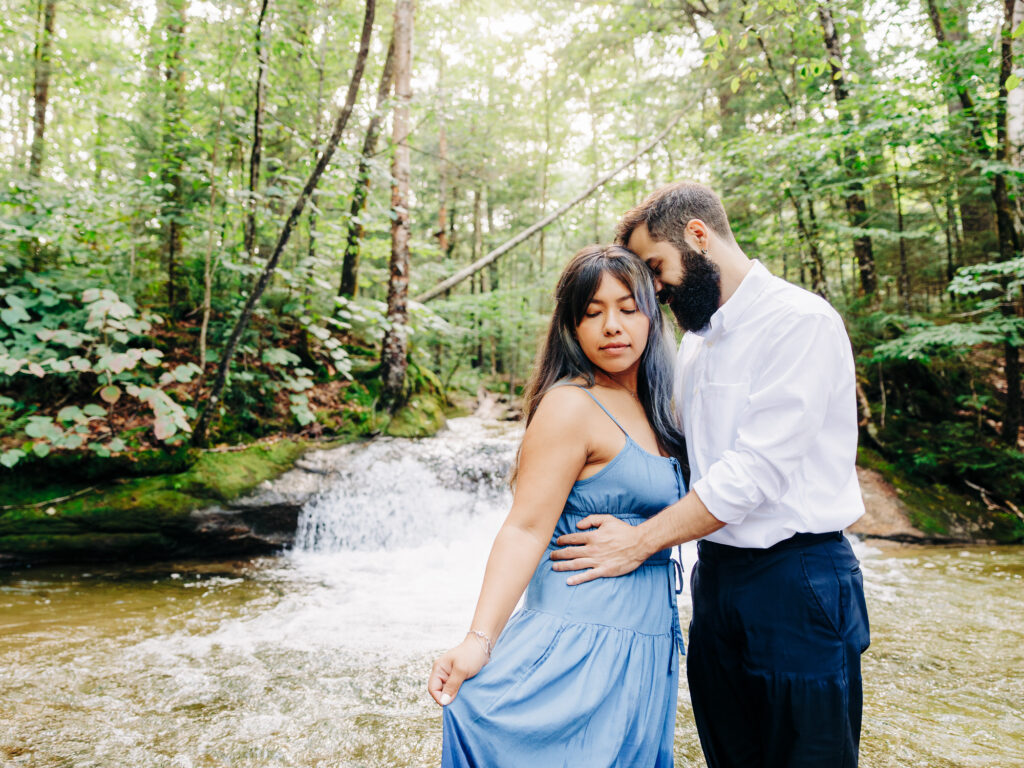 The couple stands together by a small waterfall in the woods, the woman in a light blue dress, the man in rolled-up pants and a white shirt. They share a peaceful, intimate moment, with lush greenery all around them.