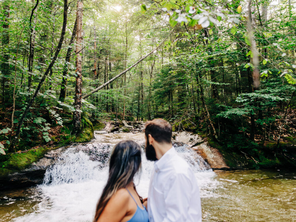 The couple stands by the edge of a stream, their backs to the camera, looking out at the flowing water and dense trees surrounding them, lost in the tranquility of nature.