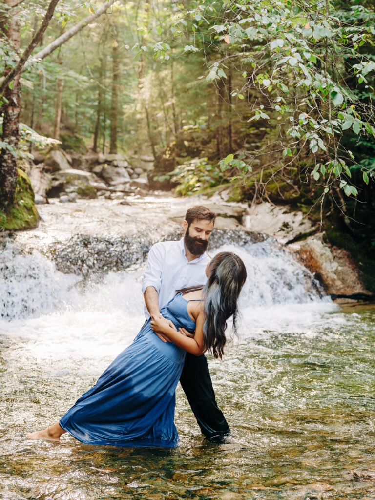 A romantic moment captured as the man dips the woman in the shallow waters of a creek, her blue dress flowing around her legs. Behind them, a small waterfall cascades down over rocks, with the soft sunlight filtering through the trees, adding a magical glow to the serene forest scene.