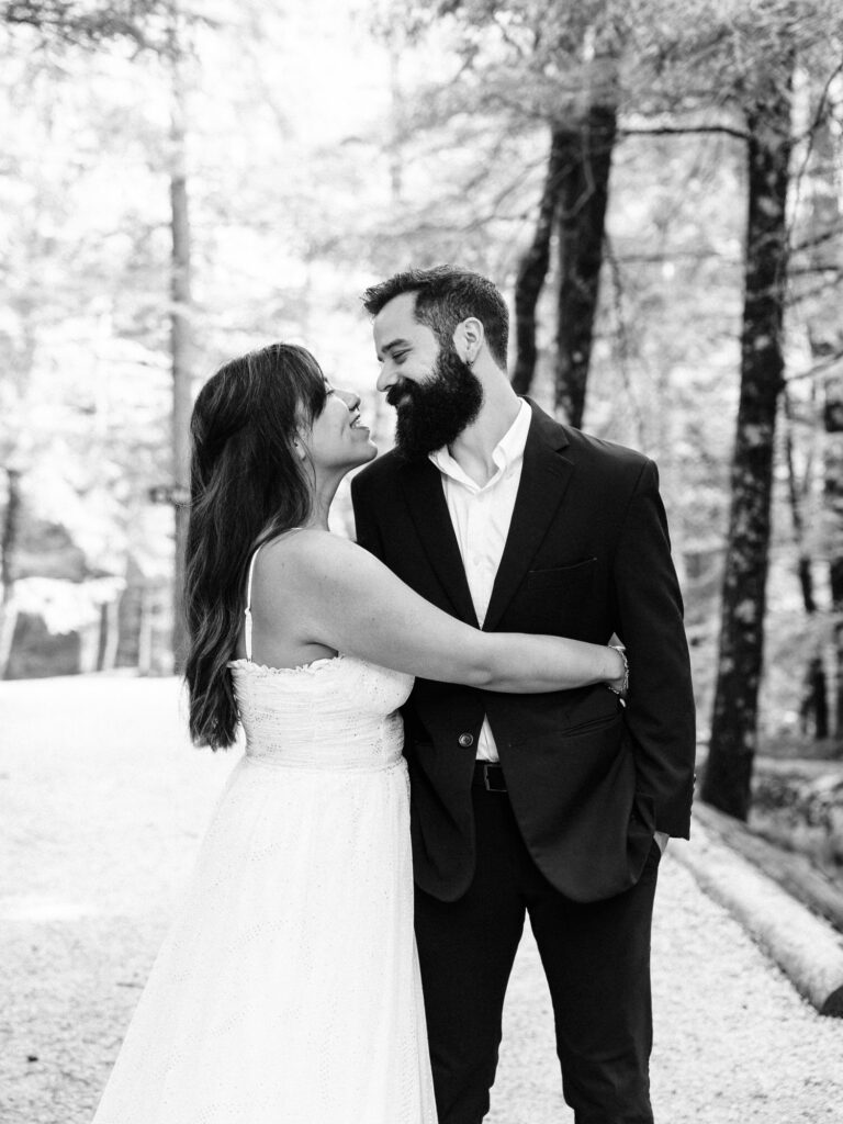 A black and white image of the couple embracing on a forest path. The woman, in a white dress, smiles as she leans in close to the man, who is wearing a dark suit. The wooded background adds a romantic, timeless feel.