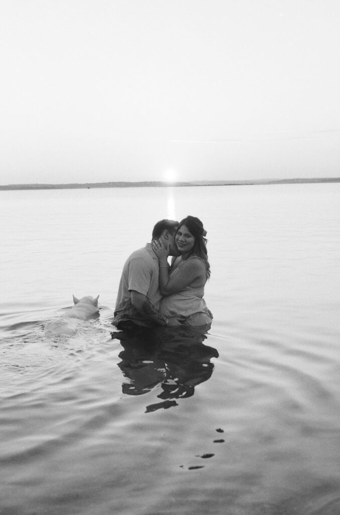 A black-and-white photo of the couple standing in the shallow water of Canyon Lake during sunset, embracing closely while their dog swims nearby.