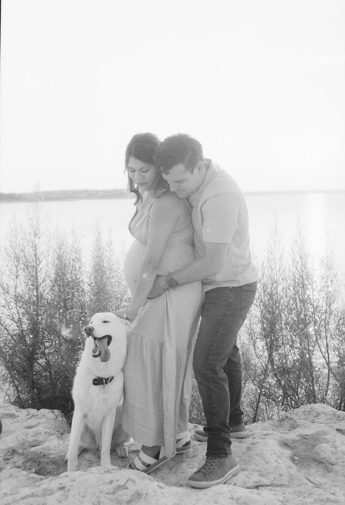 A black-and-white photo of the couple standing on the shore of Canyon Lake, with their dog happily panting beside them as they share a quiet moment together.