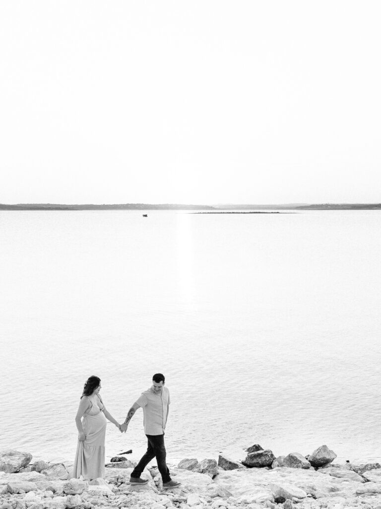 A black-and-white version of the couple walking along the shore of Canyon Lake, holding hands. The calm water and distant horizon add to the serenity of the moment.