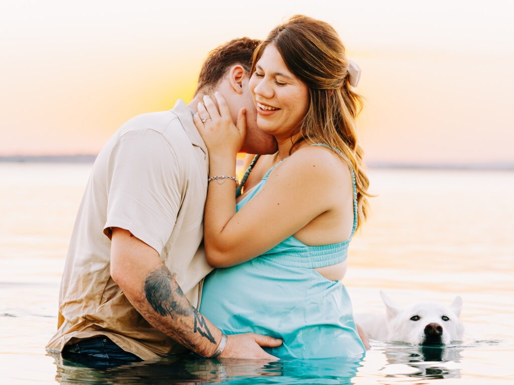 The couple standing waist-deep in Canyon Lake at sunset, with the man gently kissing the woman’s neck while cradling her pregnant belly, and their white dog floating nearby in the water.