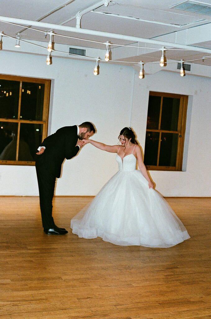 A groom in a black suit gently kisses the hand of a bride in a white ball gown as they dance in an empty, warmly lit room with wooden floors and large windows.
