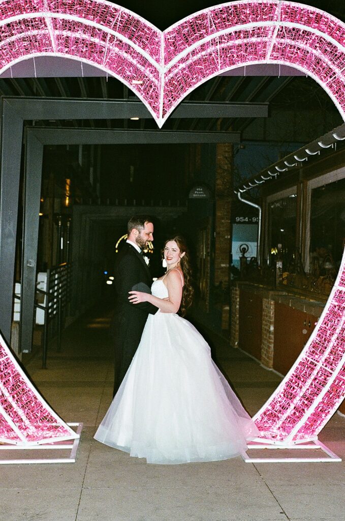 A bride and groom stand smiling under a large, pink, heart-shaped light arch at night. The bride wears a white gown and the groom wears a black suit. They hold each other and pose for the photo outdoors.