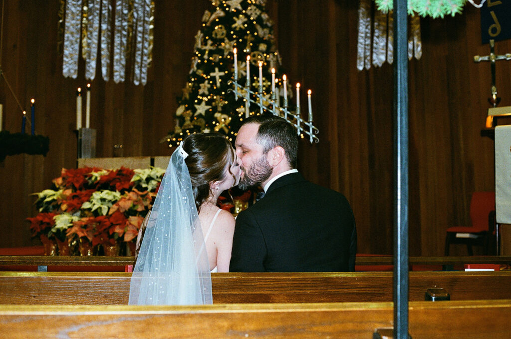 A bride and groom sit in a church pew, sharing a kiss. The bride wears a white veil, and a decorated Christmas tree with candles and poinsettias is visible in the background.