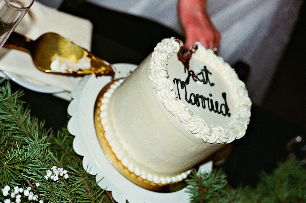 A person cuts a white frosted cake with Just Married written on top, surrounded by greenery and white flowers on a table.