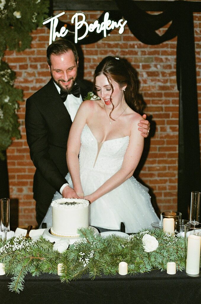 A bride and groom smile as they cut a white wedding cake together. They stand behind a decorated table with greenery, candles, and flowers. A neon sign reading The Borders hangs behind them against a brick wall.
