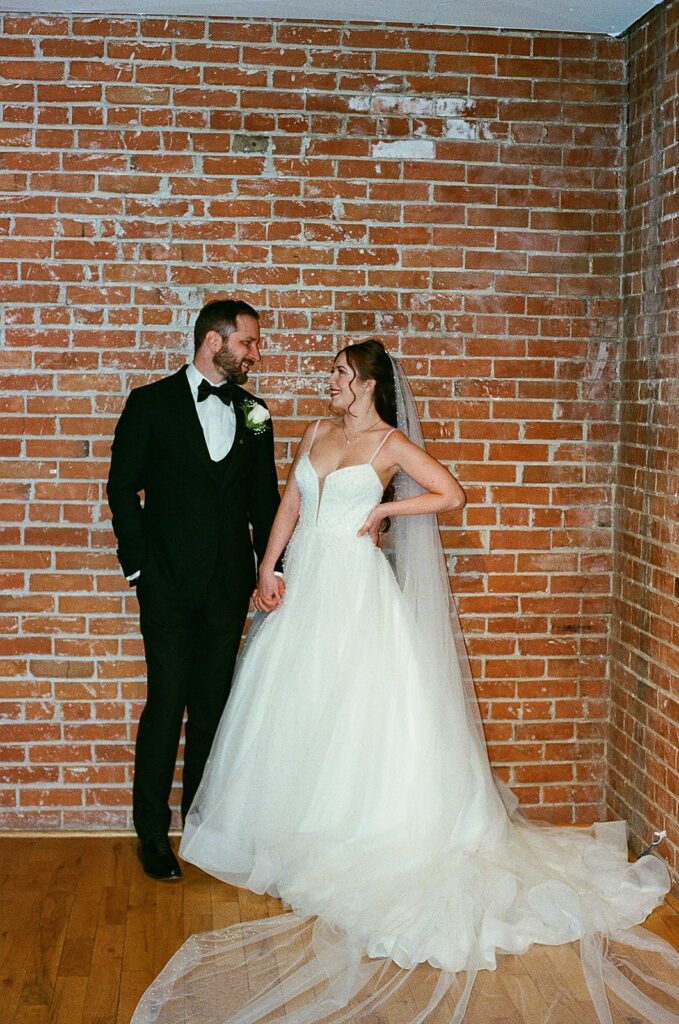 A bride in a white gown and veil holds hands and smiles at a groom in a black tuxedo as they stand together in front of a brick wall.