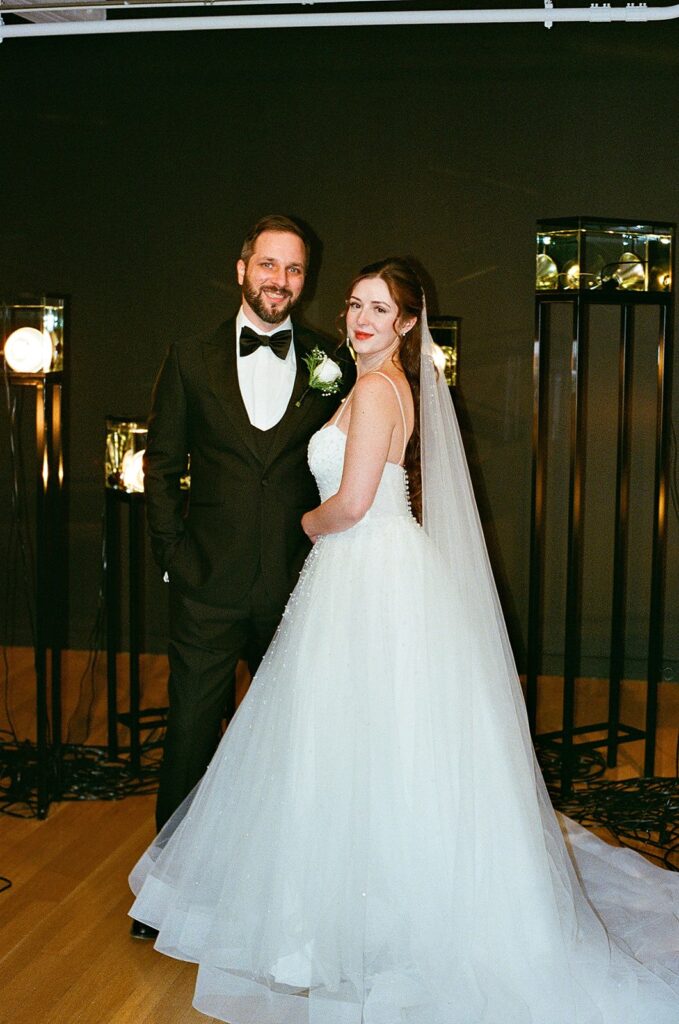 A bride in a white gown and veil stands next to a groom in a black tuxedo, both smiling and posing for a photo indoors with soft lighting and decorative lamps in the background.