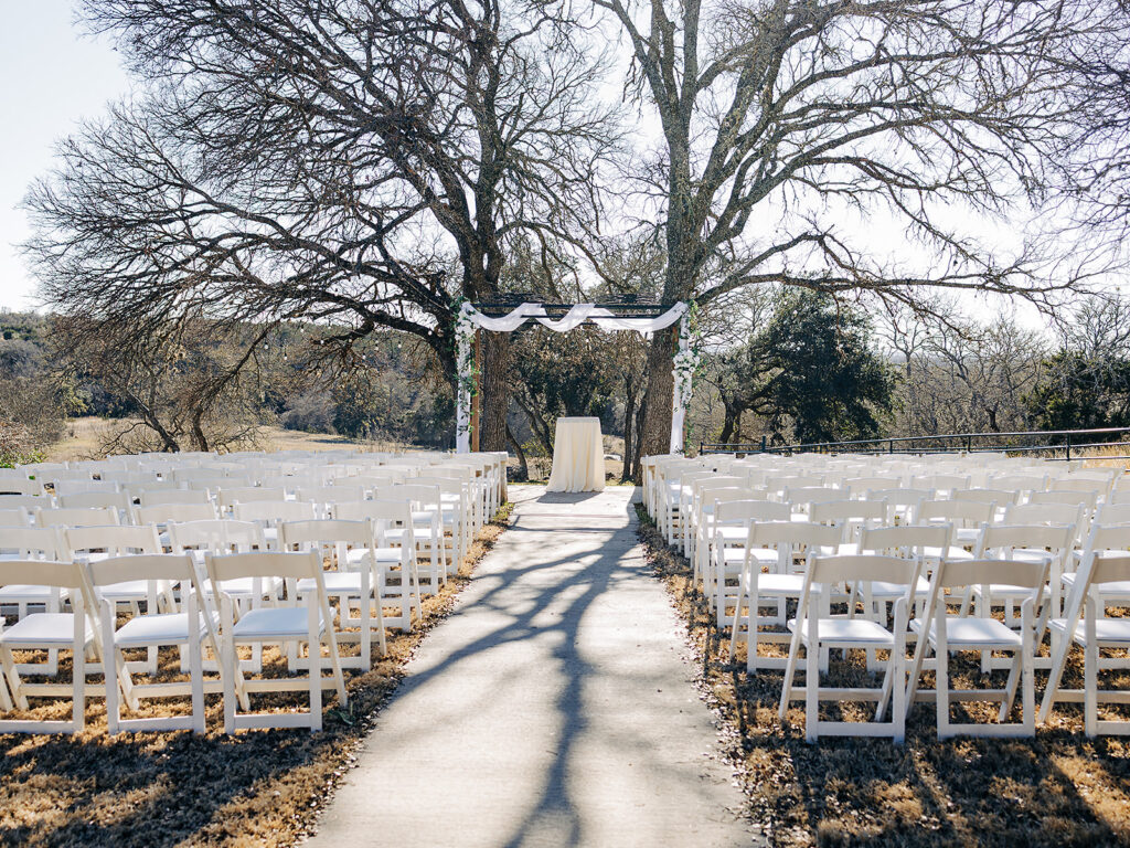 Rows of white chairs line a walkway leading to a simple outdoor wedding altar under two large leafless trees, with white fabric decorations and a sunlit natural background.