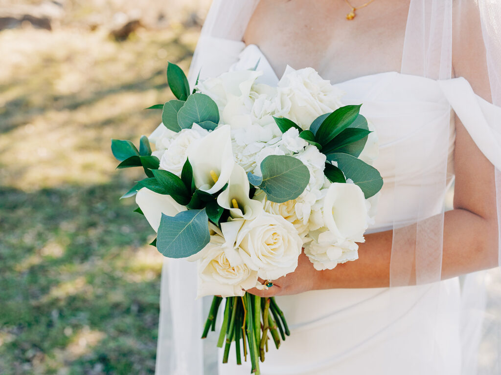 A bride in a white dress holding a bouquet of white roses, calla lilies, hydrangeas, and green leaves. She stands outdoors with a blurred grassy background.