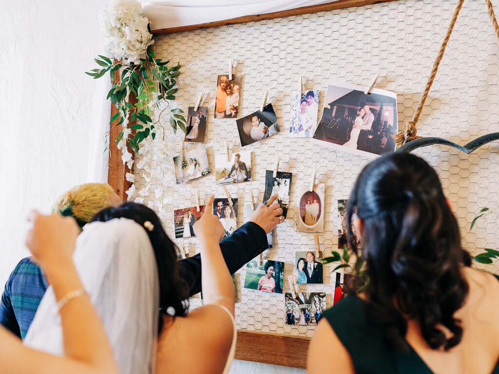 A bride and two other people look at and point to a display of old photographs clipped to a wire mesh board, decorated with flowers and greenery at a wedding.